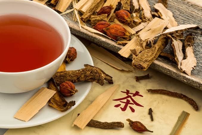 一杯沖泡好的中藥茶飲，旁邊放有多種乾燥中藥材，包括根莖、樹皮、乾果及丁香，襯以寫有「茶」字的紙箋。
